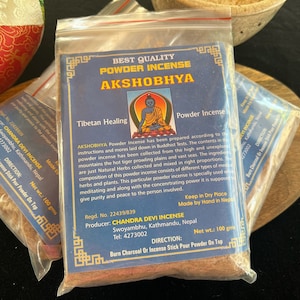 May include: A blue and white packet of Tibetan Healing Powder Incense labelled "AKSHOBHYA" with a picture of a Buddha. The packet is made by Chandra Devi Incense in Kathmandu, Nepal. The packet contains 100 grams of incense powder.