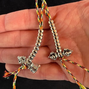 Könnte beinhalten: Zwei silberfarbene Gebetsketten mit zylindrischem Design und dekorativen Anhängern. Die Perlen sind auf farbenfrohen Schnüren mit orangefarbenen, gelben, grünen und weißen Fäden aufgefädelt. Im Hintergrund eine Hand.