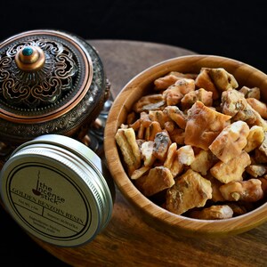 May include: A small tin of golden benzoin resin with the label "The Incense Stick Golden Benzoin Resin" in brown text on a beige background. The tin is sitting on a wooden surface next to a wooden bowl filled with golden benzoin resin chunks.
