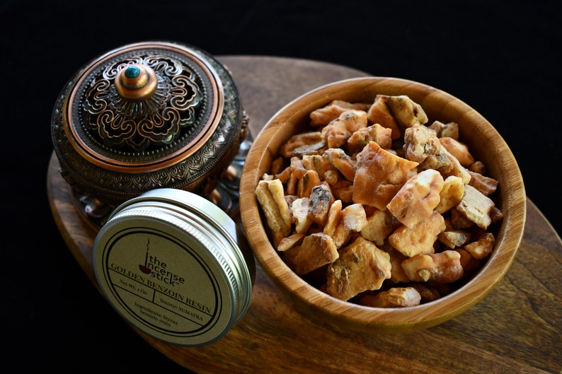 May include: A small tin of golden benzoin resin with the label "The Incense Stick Golden Benzoin Resin" in brown text on a beige background. The tin is sitting on a wooden surface next to a wooden bowl filled with golden benzoin resin chunks.