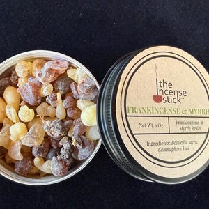 Puede incluir: Un bote de plata abierto lleno de resina de incienso y mirra, junto a un bote cerrado con una etiqueta que dice "FRANKINCENSE & MYRRH". La etiqueta también indica "Peso neto 28g" y enumera los ingredientes.