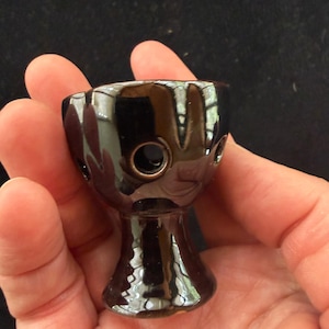 May include: A small, black ceramic object with a glossy finish. It has a rounded bowl shape with three small holes and a stem. The object is held in a hand against a dark background.