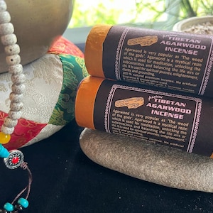 May include: Two orange and black incense sticks with the text "Tibetan Agarwood Incense" and a description of the benefits of agarwood incense. The incense is 100% natural and contains no artificial fragrance or preservatives.