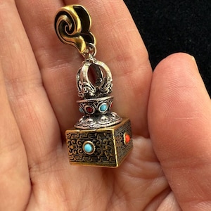 May include: A detailed pendant with a gold-colored top and a silver and gold-colored body. The pendant features turquoise and red accents. The square base has an ornate design. The pendant is held in a hand.