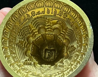May include: A gold-colored metal mold with intricate designs and a raised inscription around the rim. The mold is shaped like a bowl with a rounded bottom and a raised center.