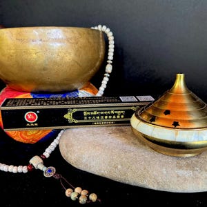 May include: A brass singing bowl with a wooden striker, a box of incense, a decorative brass container with star cutouts, and a beaded mala necklace. The items are arranged on a stone and colorful cushion.