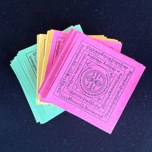 May include: Stacks of colorful prayer cards in green, yellow, and pink. Each card features intricate black text and a central circular design. The cards are arranged in a fan shape, with the pink cards at the top.