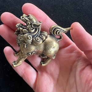 Puede incluir: Una pequeña estatua de león decorativa de color dorado, detallada, con cola enroscada. La estatua es de metal y tiene un diseño chino tradicional.