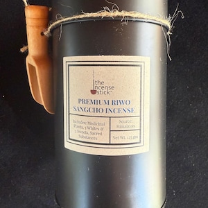 Puede incluir: Un recipiente cilíndrico negro de "Premium Riwo Sangcho Incense" con una cuchara de madera adjunta. La etiqueta incluye texto sobre ingredientes y origen. El recipiente está atado con hilo y tiene un peso neto de 125 gramos.
