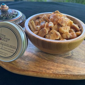 May include: A small tin container with a brown label that reads "the incense stick GOLDEN BENZOIN RESIN Net Wt. 1 Oz Source: SUMATRA Ingredients: Styrax benzoin resin" is next to a wooden bowl filled with golden brown resin chunks.