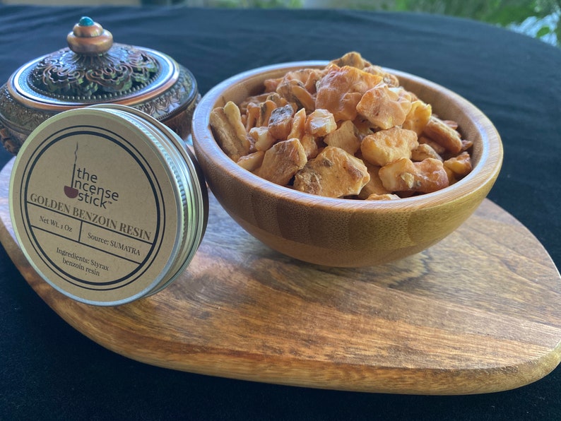 May include: A small tin container with a brown label that reads "the incense stick GOLDEN BENZOIN RESIN Net Wt. 1 Oz Source: SUMATRA Ingredients: Styrax benzoin resin" is next to a wooden bowl filled with golden brown resin chunks.