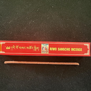 May include: A red rectangular box of Riwo Sangcho Incense with gold lettering and a small image of a deity. A single incense stick is next to the box. The background is dark.