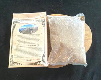 Può includere: Un sacchetto di carta marrone con una scena di montagna e testo in tibetano e inglese. Il testo recita "The Sacred Incense Powder of Lapchi". Il sacchetto contiene un sacchetto di plastica trasparente pieno di polvere marrone. Il sacchetto è appoggiato su una superficie di legno.