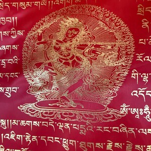 May include: A red fabric prayer flag with gold lettering and a gold image of a deity in the center. The flag features a circular design with a deity in the center, surrounded by a mandala-like pattern. The flag is decorated with Tibetan script.