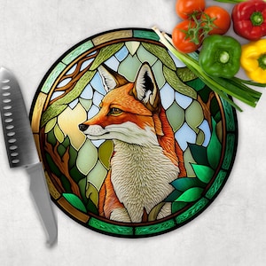 May include: A round glass cutting board with a stained glass design of a fox in a forest setting. The fox is orange and white with green leaves and branches surrounding it.