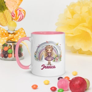 Puede incluir: Taza de cerámica blanca con borde y asa de color rosa. La taza presenta una ilustración colorida de un hada con alas de pie frente a un arcoíris. El texto "Jessica" está impreso en rosa debajo de la ilustración.