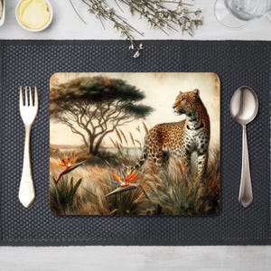 Puede incluir: Mantel individual rectangular con una ilustración vintage de un leopardo en un paisaje de sabana, con un árbol y flores de ave del paraíso. Un tenedor, una cuchara y un mantel individual gris oscuro rodean la imagen.