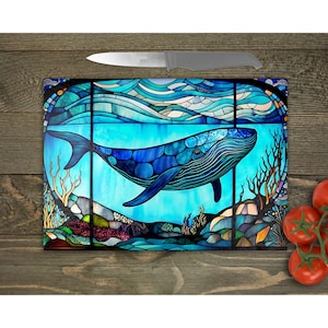 May include: A stained glass style chopping board featuring a blue whale swimming in a turquoise ocean. The whale is surrounded by coral and seaweed. The chopping board is made of tempered glass and is dishwasher safe.
