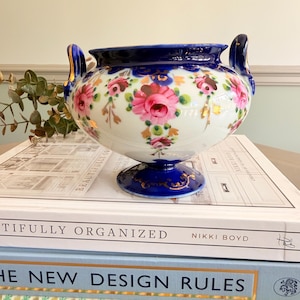 May include: A porcelain bowl with a white base, featuring hand-painted pink roses and green foliage. The bowl has a deep blue rim, a matching blue pedestal base, and two ornate blue handles with gold accents. The design includes gold leaf details.