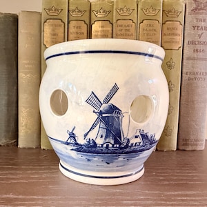 May include: A cream-colored ceramic vessel featuring a blue hand-painted windmill scene. The round container has a wide opening at the top, a flared base, and two circular cutouts on the sides. The surface displays a subtle crackle glaze finish.