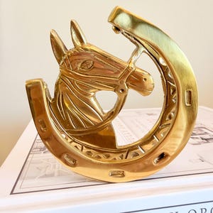 May include: A polished brass sculpture featuring a horse head centered within a horseshoe. The horse is depicted with a bridle and detailed facial features. The horseshoe has a smooth outer edge and a textured inner design with visible nail holes.