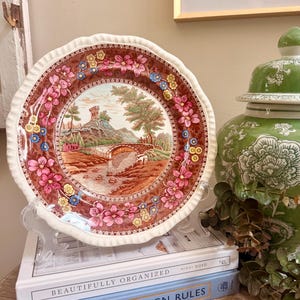 May include: A round ceramic plate with a scalloped edge featuring a central landscape illustration of a stone bridge, a river, and a castle on a hill. The border is decorated with a brown background and a pattern of pink and yellow flowers.