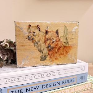 May include: A rectangular wooden block featuring a distressed, weathered finish with a painted illustration of two Cairn Terriers. One dog is light tan and the other is a darker reddish-brown, both depicted with textured, painterly brushstrokes.