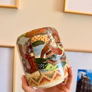 May include: A cylindrical ceramic cup featuring a hand-painted scene of a figure in traditional armor. The design incorporates intricate patterns in shades of brown, red, green, and gold, accented with raised white dots and a geometric border.