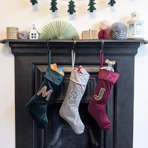 May include: Three velvet Christmas stockings hang from a black fireplace. The stockings are dark green, grey, and burgundy, each with a floral initial. A small toy mouse peeks out of the burgundy stocking. A garland of green felt trees hangs above.