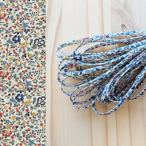 Könnte beinhalten: Nahaufnahme von Stoff mit Blumenmuster und einem Bündel passender Kordel. Der Stoff hat einen cremefarbenen Hintergrund mit kleinen roten, blauen und gelben Blumen und grünen Blättern. Die Kordel hat ein ähnliches Blumenmuster. Auf einer hellen Holzoberfläche.