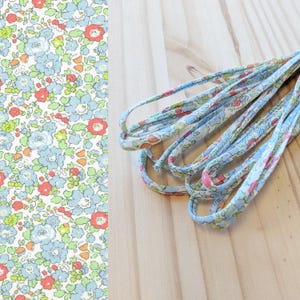 Op de afbeelding: Close-up van bloemenstof en bijpassend koord. De stof heeft een patroon van blauwe, rode en groene bloemen op een witte achtergrond. Het koord is lichtblauw met een vergelijkbaar bloemenpatroon. De items liggen op een licht houten oppervlak.