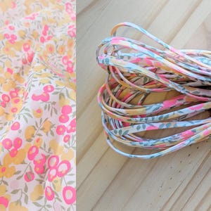 Puede incluir: Tela floral y cinta enrollada. La tela presenta un fondo blanco con estampados florales rosas y amarillos. La cinta es una mezcla de estampados florales rosas, amarillos y azules. La cinta está enrollada sobre una superficie de madera.