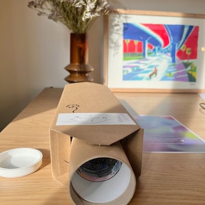 Peut inclure: Un tirage d'art cylindrique dans une bo&icirc;te en carton avec un couvercle blanc. L'impression est visible au bout du tube, avec une illustration color&eacute;e. La bo&icirc;te a une &eacute;tiquette blanche avec un petit dessin. Un vase de fleurs s&eacute;ch&eacute;es et une &oelig;uvre encadr&eacute;e sont en arri&egrave;re-plan.