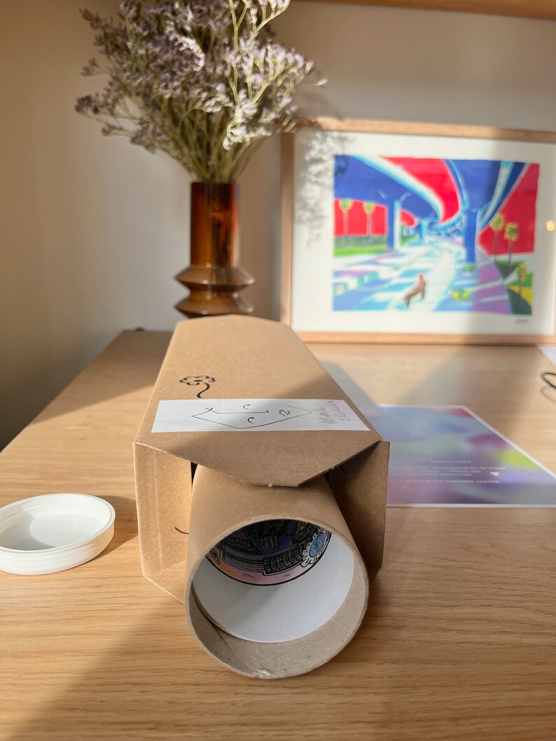 Puede incluir: Un tubo de cart&oacute;n marr&oacute;n con una etiqueta blanca y una impresi&oacute;n enrollada en su interior. El tubo est&aacute; sobre una mesa de madera con una tapa blanca y una pintura enmarcada en el fondo.