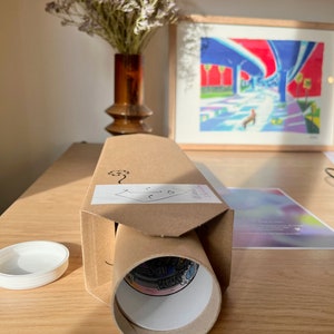 Puede incluir: Un tubo de cart&oacute;n marr&oacute;n con una etiqueta blanca y una impresi&oacute;n enrollada en su interior. El tubo est&aacute; sobre una mesa de madera con una tapa blanca y una pintura enmarcada en el fondo.