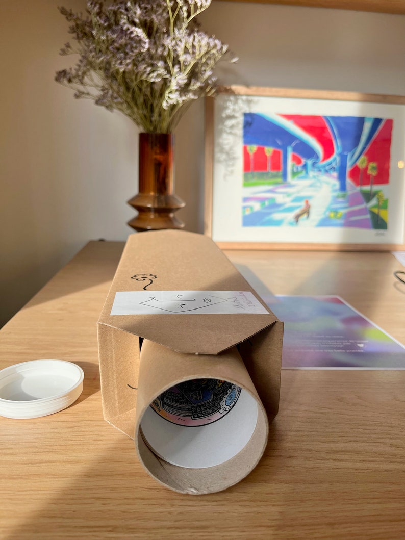 Puede incluir: Un tubo de cart&oacute;n marr&oacute;n con una etiqueta blanca y una impresi&oacute;n enrollada en su interior. El tubo est&aacute; sobre una mesa de madera con una tapa blanca y una pintura enmarcada en el fondo.