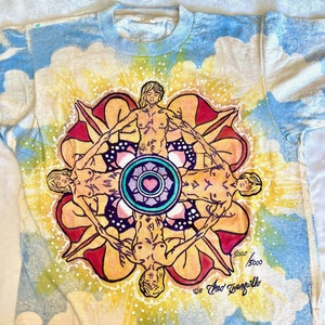 May include: A tie-dye t-shirt with a circular design featuring four figures in a mandala-like pattern. The figures are nude and have long hair. The design is in shades of yellow, orange, red, blue, and green. The shirt has a white background and is signed "Fred  Tanguillo" and "5000/5000".