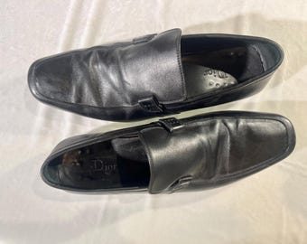 Könnte beinhalten: Ein Paar schwarze Slipper aus Leder mit einem dezenten geprägten Logo auf der Innensohle. Die Schuhe haben eine spitze Zehenpartie und einen niedrigen Absatz.