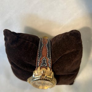 Puede incluir: Un reloj de pulsera dorado con una correa de cuero marr&oacute;n y negro. La correa presenta costuras decorativas y un dise&ntilde;o detallado. El reloj se exhibe sobre un coj&iacute;n de terciopelo marr&oacute;n oscuro.
