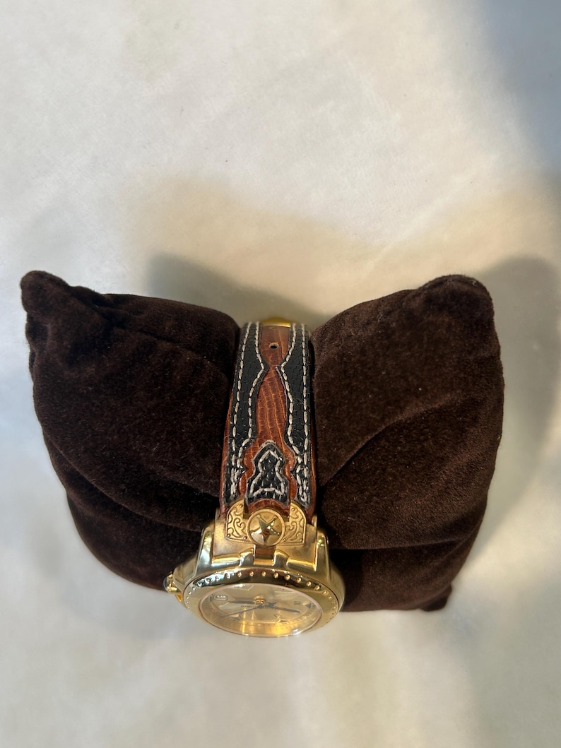 Puede incluir: Un reloj de pulsera dorado con una correa de cuero marr&oacute;n y negro. La correa presenta costuras decorativas y un dise&ntilde;o detallado. El reloj se exhibe sobre un coj&iacute;n de terciopelo marr&oacute;n oscuro.