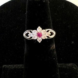 May include: A silver ring with a red gemstone surrounded by smaller clear gemstones. The ring has a delicate design with heart-shaped accents.