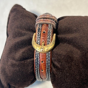 Puede incluir: Una correa de reloj de cuero marr&oacute;n con una hebilla dorada. La correa presenta un patr&oacute;n decorativo con detalles en negro, blanco y marr&oacute;n. La correa del reloj se muestra sobre un coj&iacute;n marr&oacute;n.