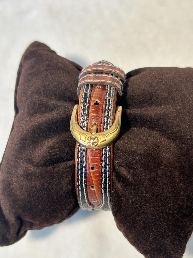 Puede incluir: Una correa de reloj de cuero marr&oacute;n con una hebilla dorada. La correa presenta un patr&oacute;n decorativo con detalles en negro, blanco y marr&oacute;n. La correa del reloj se muestra sobre un coj&iacute;n marr&oacute;n.