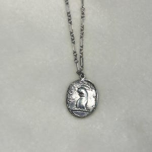 May include: A silver chain necklace with a silver oval pendant featuring a detailed image of a cat sitting down.