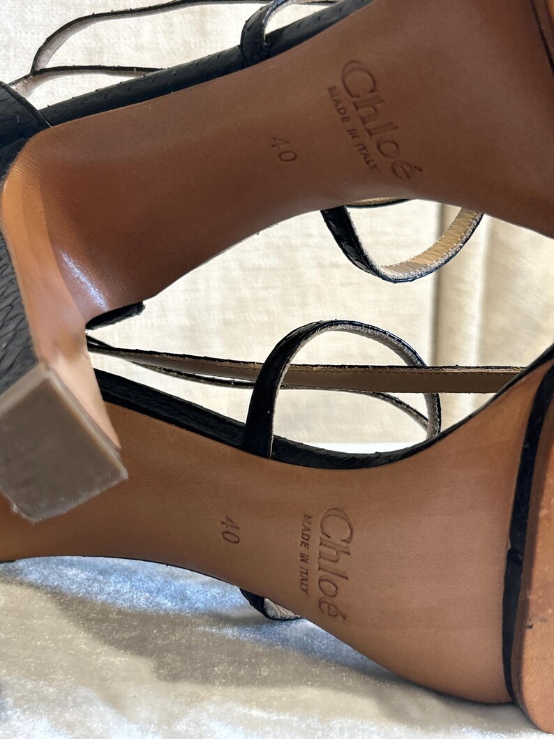 May include: Black strappy high-heeled sandals with a textured finish. The soles are tan with the brand name "Chlo&eacute;" and "Made in Italy" printed on them. The size is marked as 40.