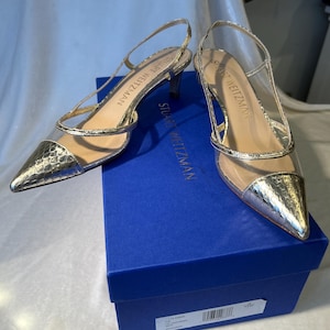 Puede incluir: Un par de tacones Stuart Weitzman dorados con correas transparentes y puntera. Los zapatos están sobre una caja azul. El área de la puntera tiene un acabado metálico texturizado. El nombre de la marca es visible.