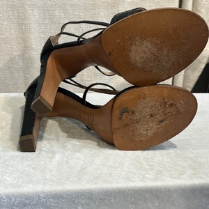 May include: Black high-heeled sandals with multiple thin straps. The sandals have a textured pattern and a square heel. The soles are brown and show signs of wear. The brand name "Chloe" is visible on the sole.