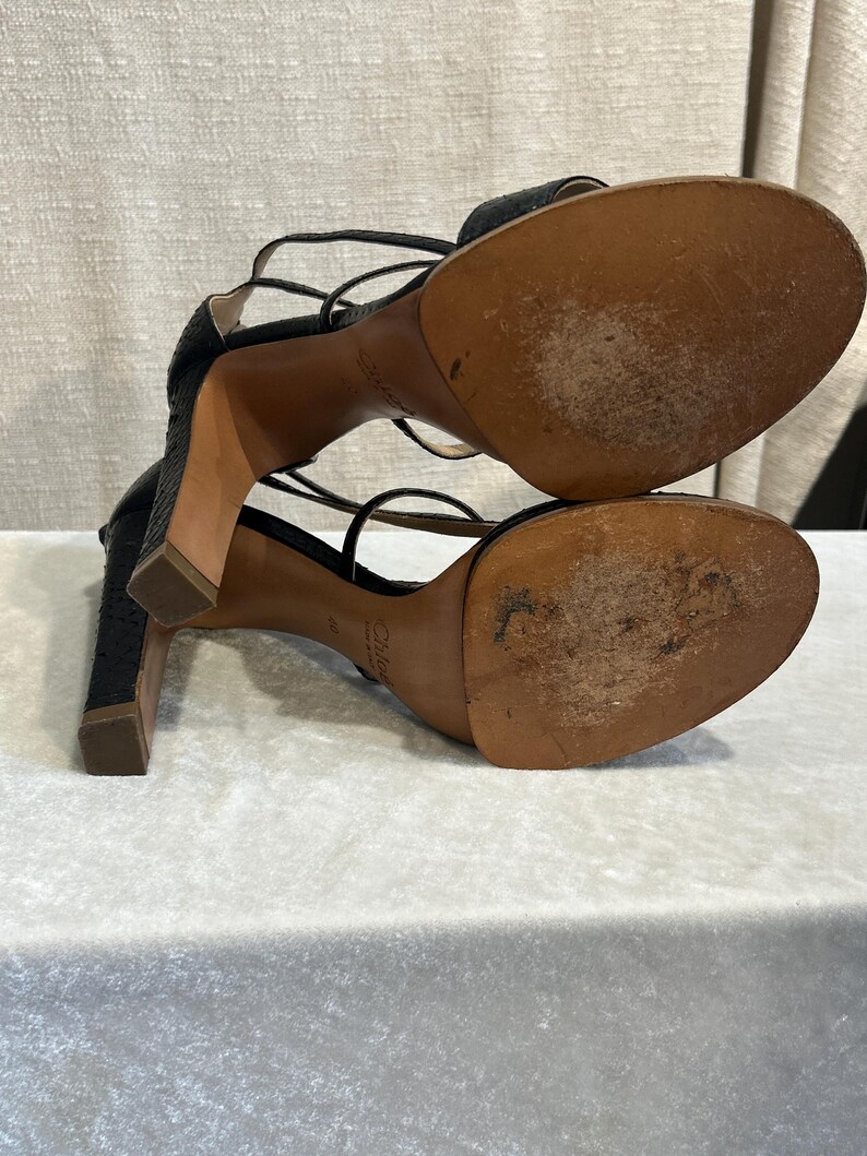 May include: Black high-heeled sandals with multiple thin straps. The sandals have a textured pattern and a square heel. The soles are brown and show signs of wear. The brand name "Chloe" is visible on the sole.