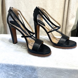 May include: A pair of black high-heeled sandals with a snakeskin texture. The sandals have multiple straps, a zip closure at the heel, and a brown sole. The heels are approximately 4 inches (10 cm) high.