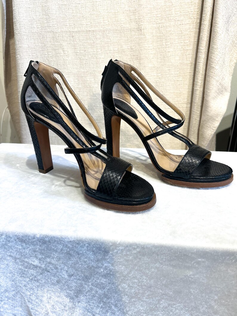 May include: A pair of black high-heeled sandals with a snakeskin texture. The sandals have multiple straps, a zip closure at the heel, and a brown sole. The heels are approximately 4 inches (10 cm) high.
